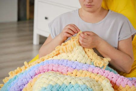 A young woman knits a warm bright plaid from multi-colored terry looped yarn with her hands. A cozy...