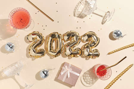 beige festive background for new year or christmas with pink drinks punch or champagne and foil gold balloons in the shape of 2022, golden confetti, disco ball and giftsの写真素材
