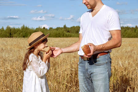 Young man breaks off an appetizing piece of bread for his little daughter in a white dress in a rye field sunny dayの写真素材