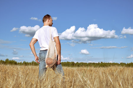 A young man stands on a sunny day in a rye field. A man holds an ecological shopping bag, a shopping bag with breadの写真素材
