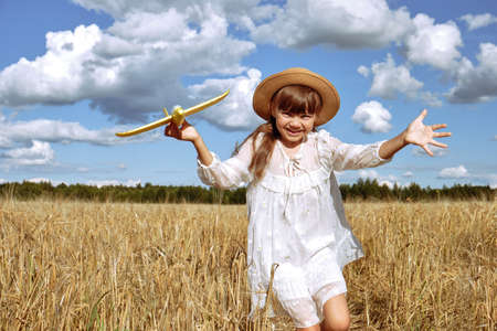A little girl in a white dress and a straw hat plays with a toy plane in a rye field inの写真素材