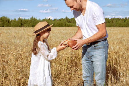Young man breaks off an appetizing piece of bread for his little daughter in a white dress in a rye field sunny dayの写真素材