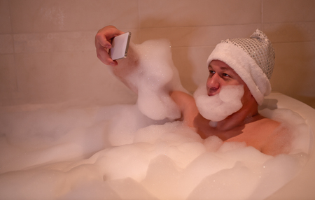 Young man sits in the bathroom, makes a beard of foam and takes a selfieの写真素材