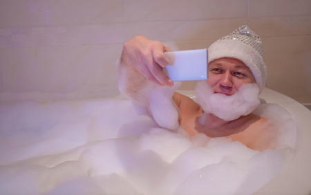 Smiling man sits in the bathroom, makes a beard of foam and takes a selfieの写真素材
