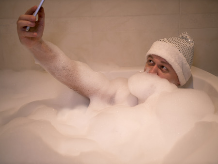 miling man sits in the bathroom, makes a beard of foam and takes a selfieの写真素材