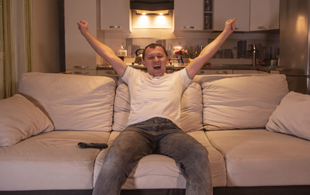 Young energetic man watching a game at home sitting on the sofa in the evening on TV, supports the football team, rejoices the goalの写真素材