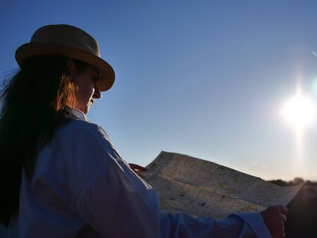 Tourist girl in a hat is considering a road map on the background of the sunrise
