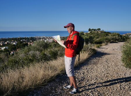 Tourist guy is standing on a hill path and holding a map, looking around