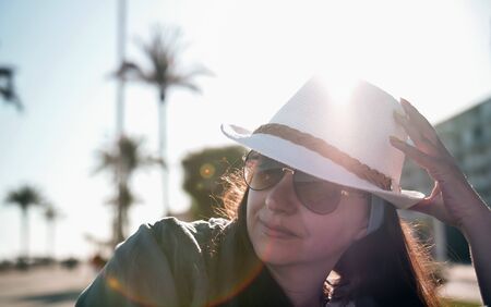 Portrait of an attractive brunette girl in a hat and sunglasses, sun above her head, sun glare