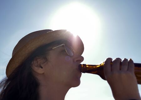 Blurred silhouette of a brunette girl in a hat drinking a drink from a glass bottle on a background of bright sunの写真素材
