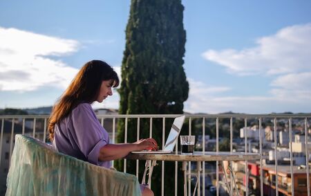 Attractive brunette girl uses her laptop while sitting on the balcony in summerの写真素材