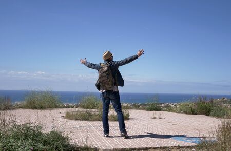 A man traveler with a backpack raised his arms high while standing on the observation deck on top of...