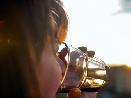 Brunette woman drinks a drink from a glass on a sunset background, close-up, sun glare