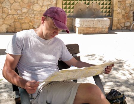 A young man in a cap sits on a street bench with a map in his handsの写真素材
