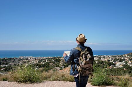 A young man with a backpack and a map of the area in his hands stands on a mountain near the city...