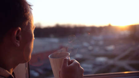 A man stands at the window and looks at the sunset, drinks tea from a white mug, close-upの写真素材