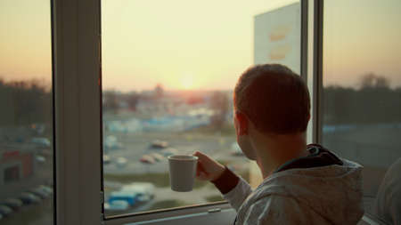 A man stands by the window and watches the sunset, drinks tea from a white mug, rear viewの写真素材