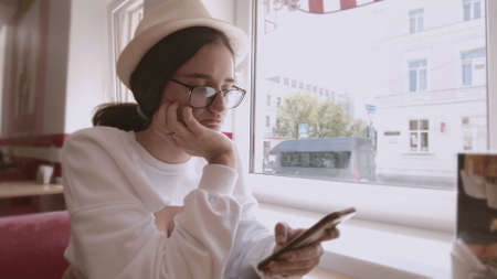 Girl in a hat and glasses is sitting in a cafe, using her cell phone, waiting for her order