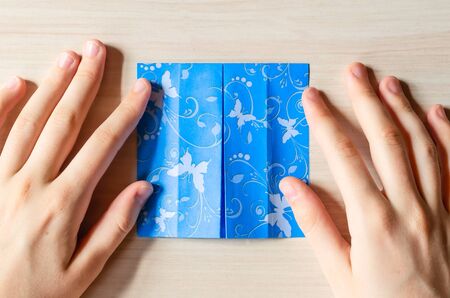 Girl's hands making origami blue dress from butterfly printed paper. Step by step instruction, step10. Mother's day, birhday, summer gift, family leisure time concept. DIY tutorial, flatlay, top view.の写真素材