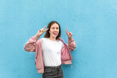 Portrait of funny caucasian teen girl in blank white t-shirt and pastel pink bomber jacket against blue city wall solid background. Emotion, generation Z, fashion, modern urban style, mockup concept.の写真素材