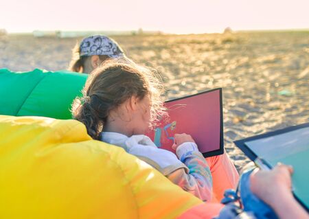 Side view of little girl sitting along other kids in colorful bag chair and drawing landscape with pastel at the beach.の写真素材