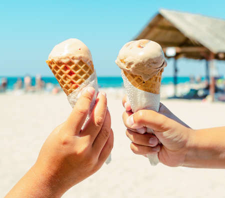 Couple kids hands holding ice cream cones at the beach background in summer vacation. Summer food, summertime joy, vacation, holidays.の写真素材