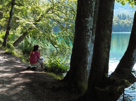 Wonderful beauty of great Nature of Germany. Mountain lake Alpsee with dreaming girl sitting on the shore. Cool shadow from tree in summer noon. Blue and green colored clean water. Moments of journey.の写真素材