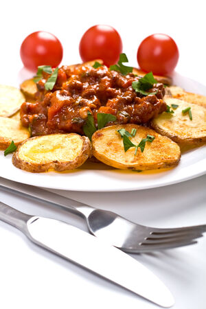 roasted country-styled potatoes with meat tomato sauce on a white plate, a fork and a knife and three cherry tomatoes at the backの写真素材