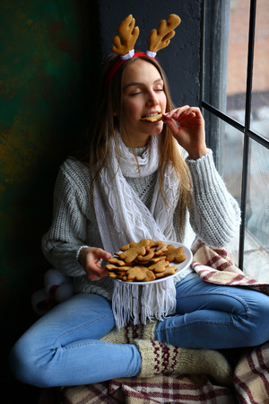 Young woman in deer horns enjoying Christmas cookies, sitting by the window with cross-leggedの写真素材