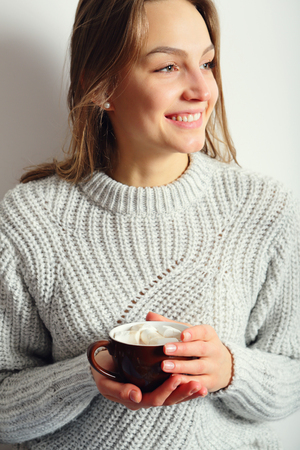 Smiling Young beautiful woman holding a mug with cocoa and marshmallow, looking awayの写真素材