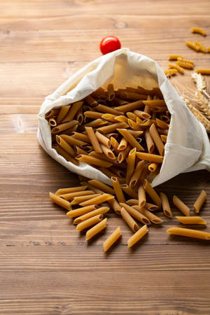 Whole grain penne pasta in white cotton bag on wooden surfaceの写真素材