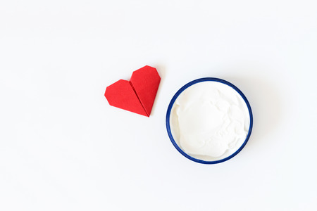 Cream for body or face in blue container on white background. Red origami heart. Caring for the body, a gift for a loved oneの写真素材