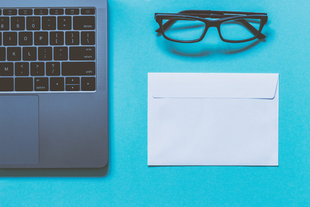 Eyeglasses, white paper envelope and laptop. Work space. Blue background. Top view.