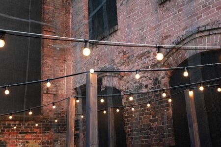 An empty courtyard lit by garlands of warm light bulbs. Old brick walls with large arched openings...