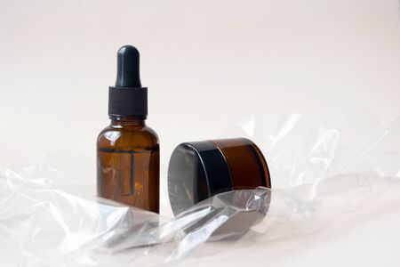Serum oil essence in brown glass bottle and cream in jar on crumpled plastic texture on light beige...