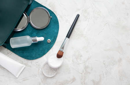 Transparent powder, makeup brush, pocket mirror, facial serum and hand cream on marble table and copy space, top view in small emerald female bag. Beauty skincare productsの写真素材