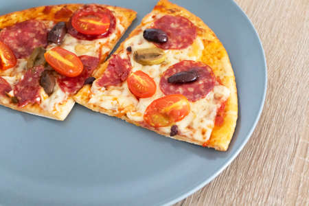 Two pieces of homemade cheese pizza with salami, olives and cherry tomato on gray plate on wooden...