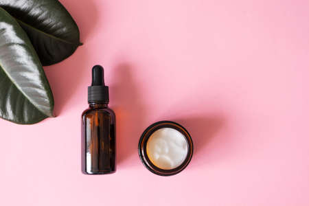 Facial serum fluid bottle with dropper and moisturizer cream on pink background and green tropical leaves. Eco organic natural beauty products for summer skin care. Dermatology and cosmetologyの写真素材