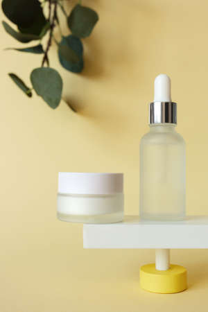 Moisturizer cream and serum in bottle standing on abstract pedestal on pastel yellow background with copy space and defocused eucalyptus leaves, front view. Skincare beauty products mockup, verticalの写真素材