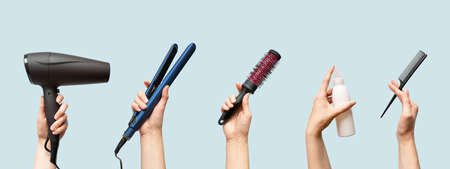 Woman hands holding hairdryer, straightener, hairbrush, tail comb and hair care essence in bottle isolated on blue background, horizontal banner formatの写真素材