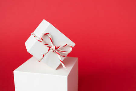Close-up Christmas gift box with striped twine cotton string balancing on cube podium on red background with copy space. Winter holidays modern layoutの写真素材