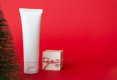 White cosmetic cream tube and gift box with red white striped twine rope on red bacground with copy space. Minimalist skincare and body care winter sale conceptの写真素材