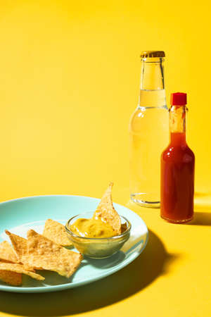 Nachos chips with cheese sauce on blue plate and soda bottle and hot sauce on yellow background. Food photo, verticalの写真素材