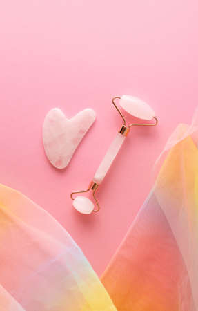 Rose quartz facial roller and gua sha on pink background with colorful rainbow organza fabric