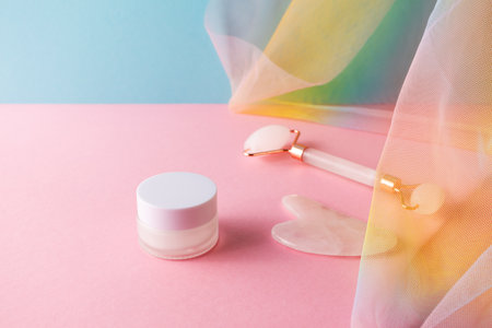 Moisture cream, quartz facial roller and gua sha for skincare massage on blue pink background colorful rainbow draperyの写真素材
