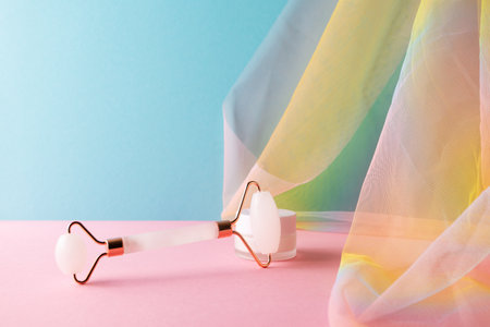 Quartz facial roller and moisture cream for skincare on blue pink background with colorful rainbow organza draperyの写真素材