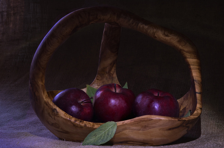 Composition with three red apples in wooden basket decorated by few green leaves on rough natural sacking material. Unfocused background.の写真素材