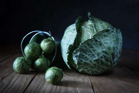 Whole heads of white cabbage and Brussels sprouts falling out of decorative metal bucket on natural wooden table. Selective focus. Rustic style. Natural evening light or moonlight.の写真素材