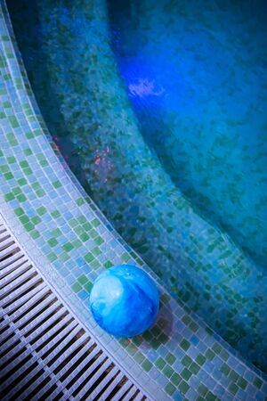 Child's blue ball is left alone in swimming pool. Stairs are visible through water where red and blue illumination is reflected. Summer atmosphere and private safety on water concepts. Copy space.の写真素材