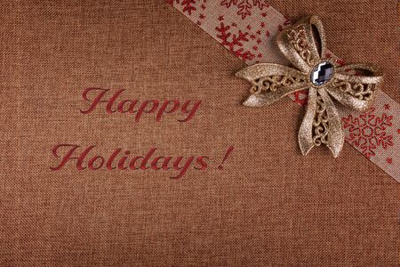 Gold glittered tied bow on beige burlap ribbon with red snowflakes festive ornament. Christmas decor is on brown natural burlap backgound. Ready to use seasonal greeting card with Happy Holidays sign.の写真素材